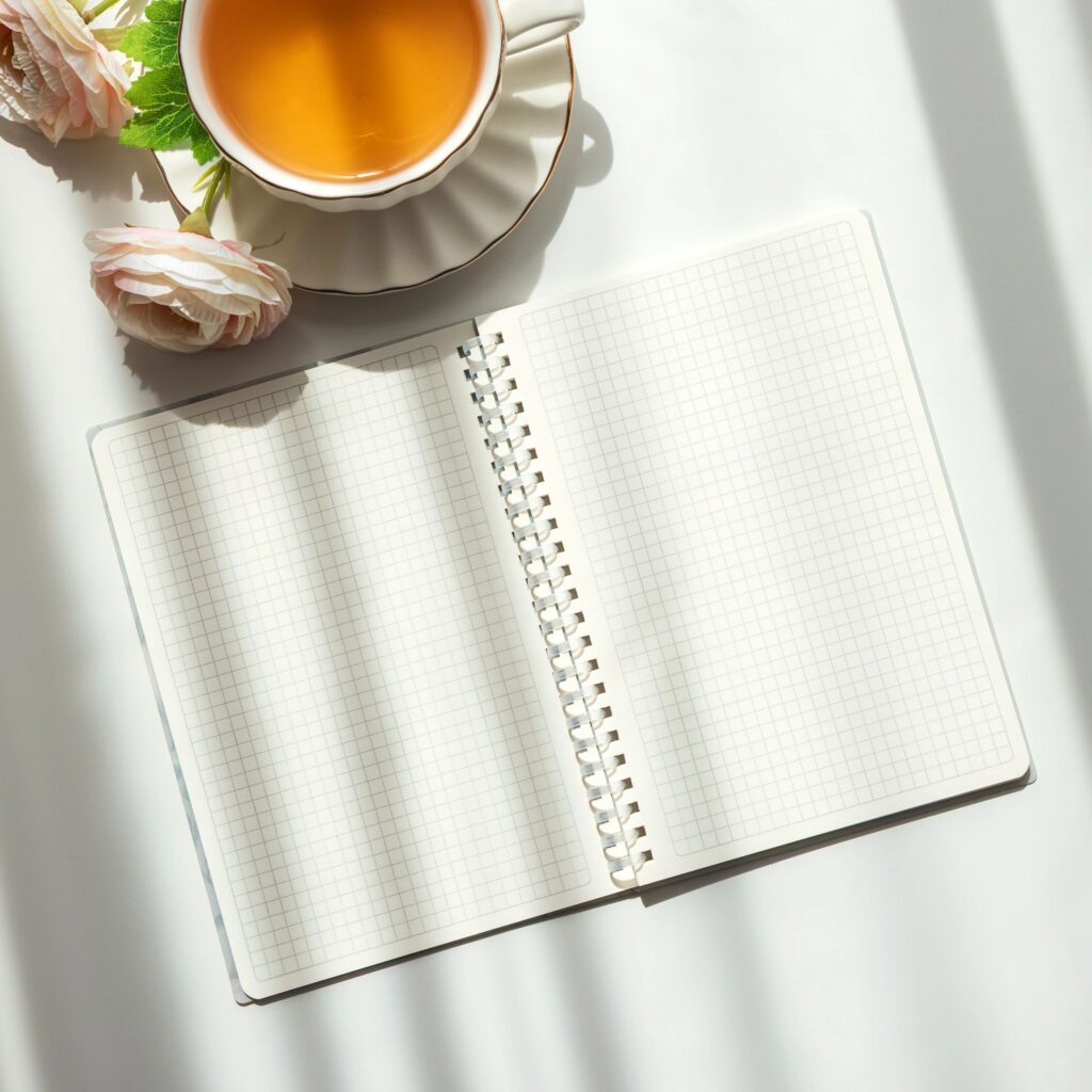 Bright lifestyle composition featuring a white teacup on a scalloped saucer, an open spiral notebook with grid paper, and a bouquet of pale pink and cream flowers.